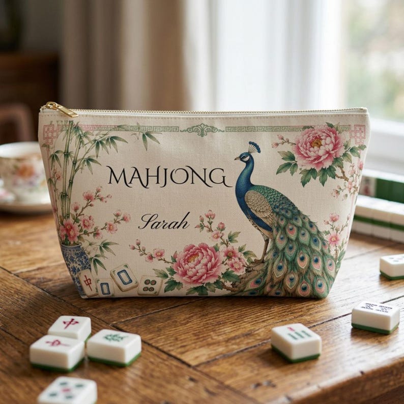 May include: A cream-colored zippered pouch featuring a printed design of a peacock, pink peony flowers, bamboo stalks, and mahjong tiles. The text "MAHJONG" and the name "Sarah" are printed in black on the front of the fabric accessory bag.