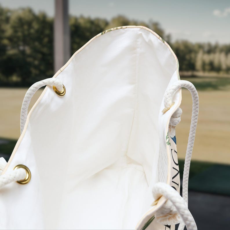 May include: A white tote bag featuring thick white rope handles attached with gold-toned metal grommets. The interior is lined with plain white fabric, and a portion of the exterior displays a floral pattern with the partial text "ING" visible.