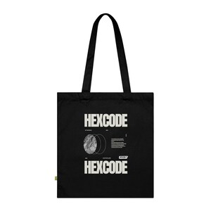 May include: Black tote bag with the word "HEXCODE" in white block letters at the top and bottom. The bag features a graphic design with a circular image and text. The tote bag has black handles.