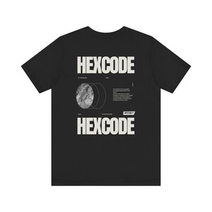 Op de afbeelding: Zwart T-shirt met een wit grafisch ontwerp met de tekst "HEXCODE" en een gestileerde afbeelding van een planeet met cirkels eromheen.
