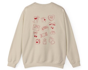 May include: Cream-colored sweatshirt with red line art illustrations. Designs include a wine glass, playing cards, a heart-shaped candy with "CRAZY 4 U", an envelope, a candy bar, bows, dice, a teddy bear, and strawberries.