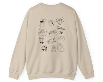 May include: A cream-colored sweatshirt featuring a collection of black line art illustrations. The drawings include a wine glass, playing cards, a heart-shaped candy with "CRAZY 4 U" text, bows, an envelope, a candy bar with "PERFECT MATCH" text, swans, dice, a teddy bear, and hearts.