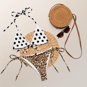 May include: A white bikini top with black polka dots and a leopard print bikini bottom are displayed with a woven straw bag, sunglasses, and a round woven mat. The bikini top has halter straps.
