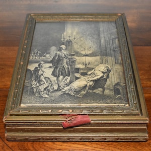 May include: A wooden box with a gold-painted finish and a hinged lid. The lid features a framed black and white print of a scene with three people. The box has a red fabric pull tab.