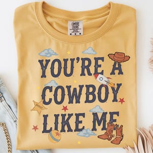 May include: A mustard yellow t-shirt featuring the dark blue text "YOU'RE A COWBOY LIKE ME" surrounded by illustrations of a brown cowboy hat, a rocket ship, a sheriff star badge, a ball, and brown cowboy boots, along with small clouds and stars.