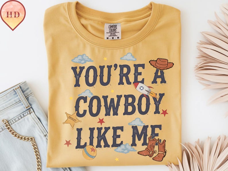 May include: A mustard yellow t-shirt featuring the dark blue text "YOU'RE A COWBOY LIKE ME" surrounded by illustrations of a brown cowboy hat, a rocket ship, a sheriff star badge, a ball, and brown cowboy boots, along with small clouds and stars.