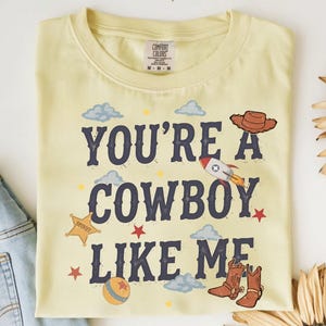 May include: A pale yellow t-shirt featuring the dark blue text YOU'RE A COWBOY LIKE ME. The design includes a brown cowboy hat, a rocket ship, a yellow sheriff star, a ball, and brown cowboy boots, surrounded by small blue clouds and red stars.