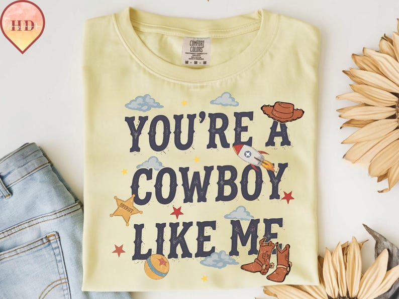 May include: A pale yellow t-shirt featuring the dark blue text YOU'RE A COWBOY LIKE ME. The design includes a brown cowboy hat, a rocket ship, a yellow sheriff star, a ball, and brown cowboy boots, surrounded by small blue clouds and red stars.