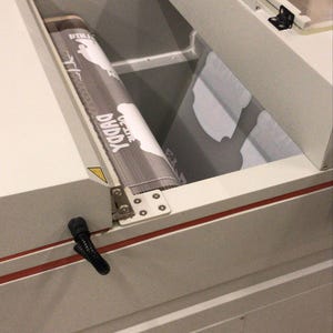 May include: A light gray industrial conveyor belt machine with a clear hinged lid. A printed film with the text "DUDDA" and "ON-LINE" is visible on the belt. The machine features a metal frame, a red accent stripe, and a lower storage drawer.