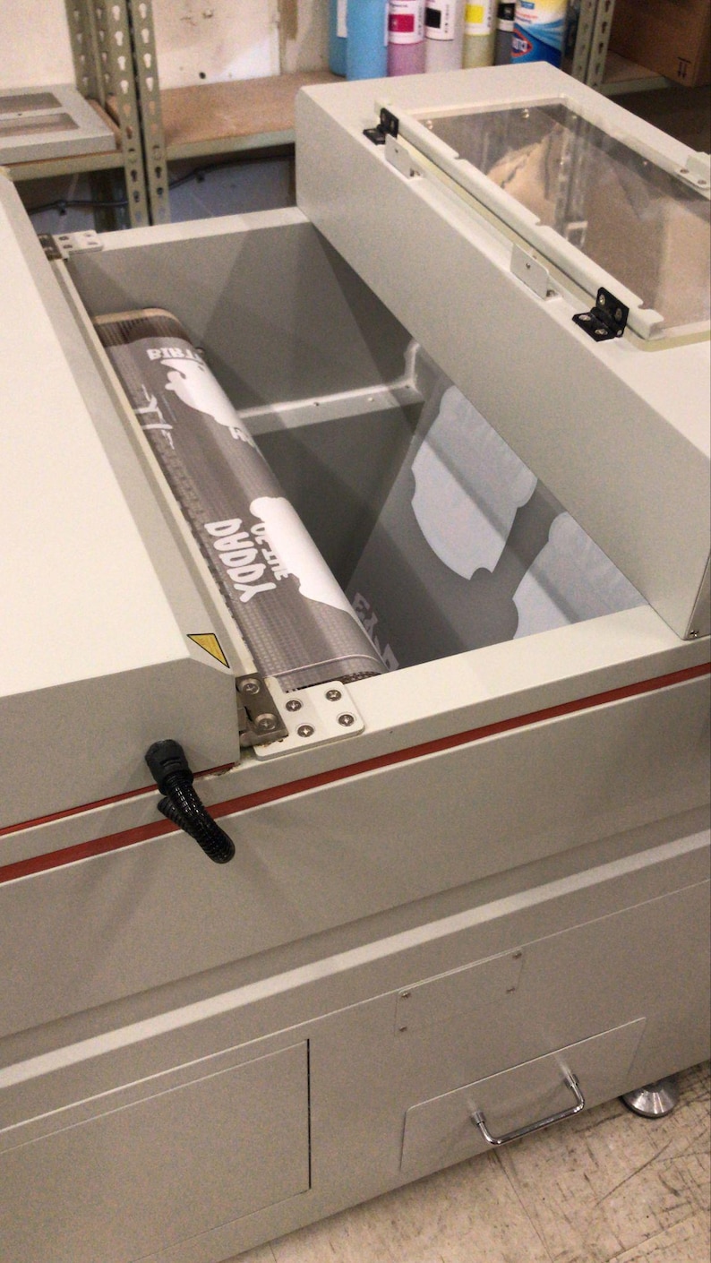 May include: A light gray industrial conveyor belt machine with a clear hinged lid. A printed film with the text "DUDDA" and "ON-LINE" is visible on the belt. The machine features a metal frame, a red accent stripe, and a lower storage drawer.