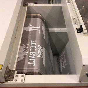 May include: A close-up of a white and gray screen printing machine with a gray fabric being printed with the words "Yoooo" and "Football" in white.
