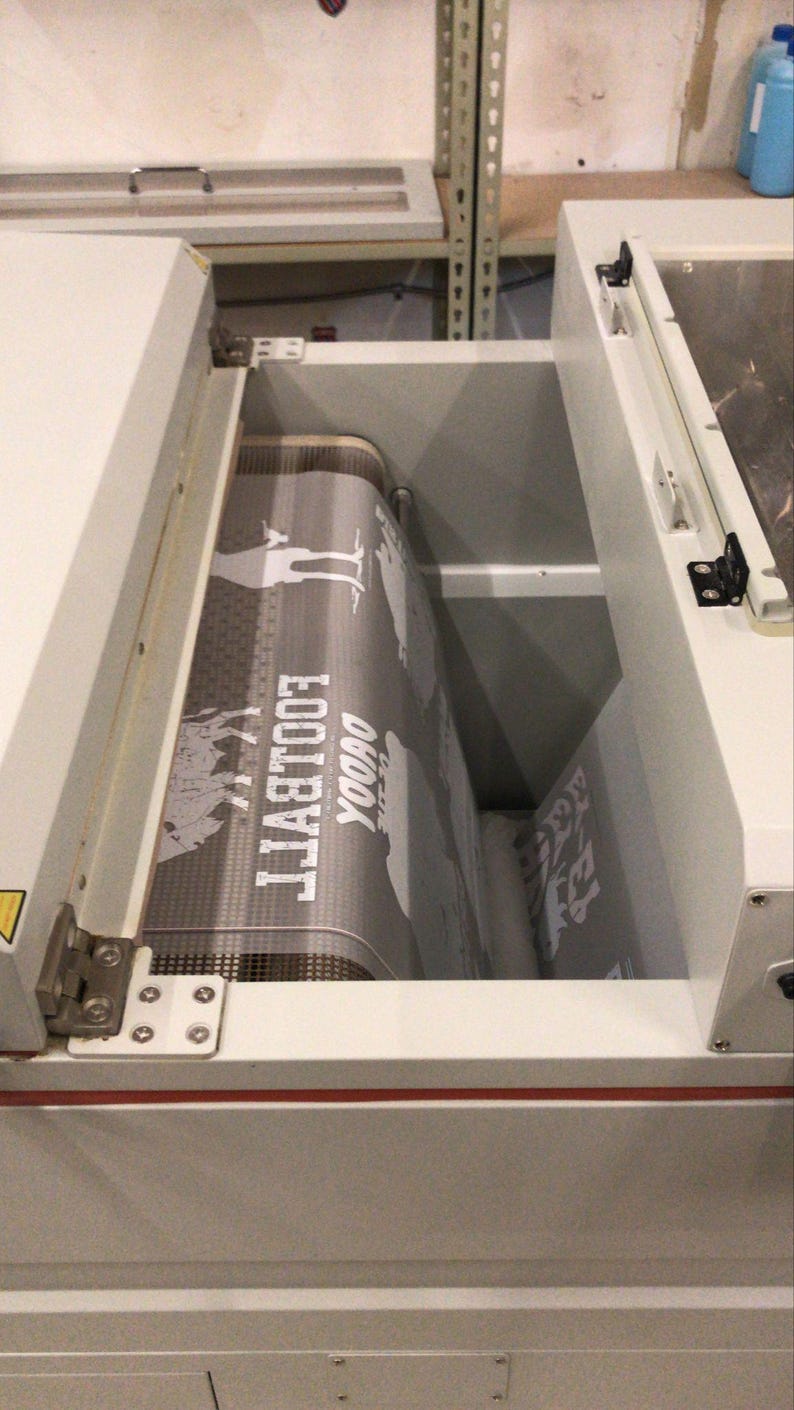 May include: A close-up of a white and gray screen printing machine with a gray fabric being printed with the words "Yoooo" and "Football" in white.
