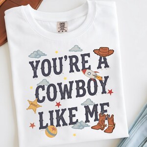 May include: A white t-shirt featuring the dark blue text "YOU'RE A COWBOY LIKE ME" surrounded by illustrations of a brown cowboy hat, a rocket ship, a yellow sheriff star, a ball, red stars, and light blue clouds on the front of the garment.