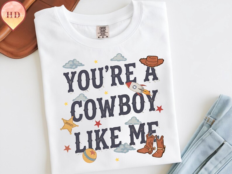 May include: A white t-shirt featuring the dark blue text "YOU'RE A COWBOY LIKE ME" surrounded by illustrations of a brown cowboy hat, a rocket ship, a yellow sheriff star, a ball, red stars, and light blue clouds on the front of the garment.