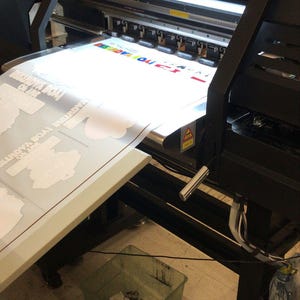 K&ouml;nnte beinhalten: Ein Gro&szlig;formatdrucker mit einem wei&szlig;en Blatt Papier, das durch die Walzen gef&uuml;hrt wird. Das Papier hat eine Karte darauf gedruckt. Im Vordergrund befindet sich ein gr&uuml;ner M&uuml;lleimer mit wei&szlig;en Papierresten und ein roter Eimer mit einem wei&szlig;en Tuch.