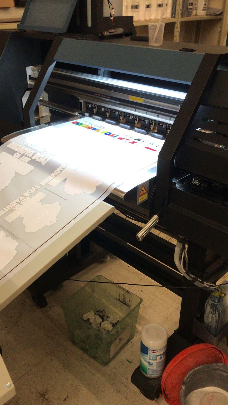 K&ouml;nnte beinhalten: Ein Gro&szlig;formatdrucker mit einem wei&szlig;en Blatt Papier, das durch die Walzen gef&uuml;hrt wird. Das Papier hat eine Karte darauf gedruckt. Im Vordergrund befindet sich ein gr&uuml;ner M&uuml;lleimer mit wei&szlig;en Papierresten und ein roter Eimer mit einem wei&szlig;en Tuch.