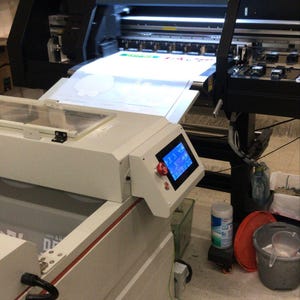 May include: A large-format printer with a white sheet of paper being fed through the machine. The printer is on a stand with a digital display and a red button. The printer is in a workshop setting with a white tiled floor.