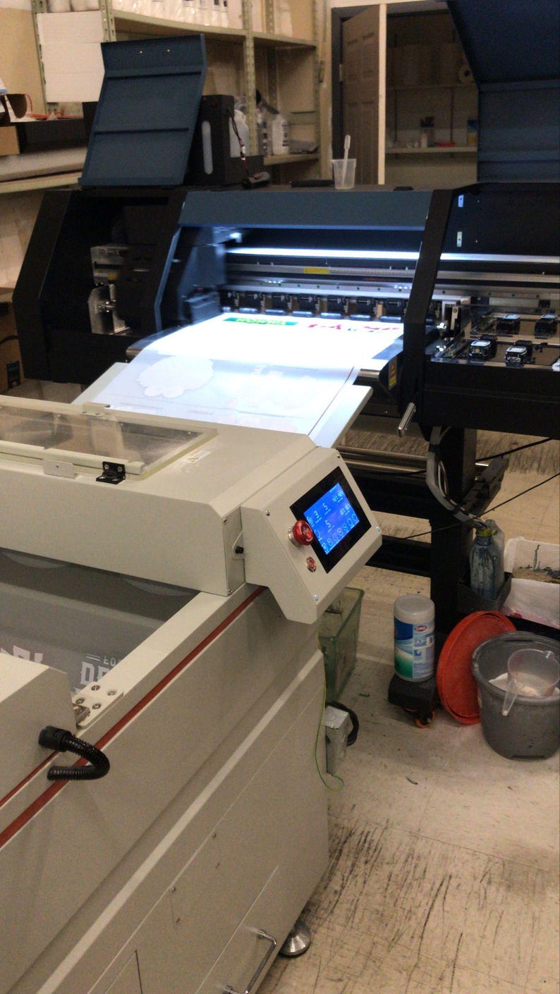 May include: A large-format printer with a white sheet of paper being fed through the machine. The printer is on a stand with a digital display and a red button. The printer is in a workshop setting with a white tiled floor.