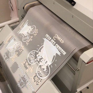 K&ouml;nnte beinhalten: Nahaufnahme einer wei&szlig;en Hei&szlig;presse mit einer durchsichtigen Plastikfolie, auf der ein Design aufgebracht ist. Das Design ist eine wei&szlig;e Silhouette einer Comicfigur mit dem Text "Sweet Feliz del Cumple" auf der Folie.