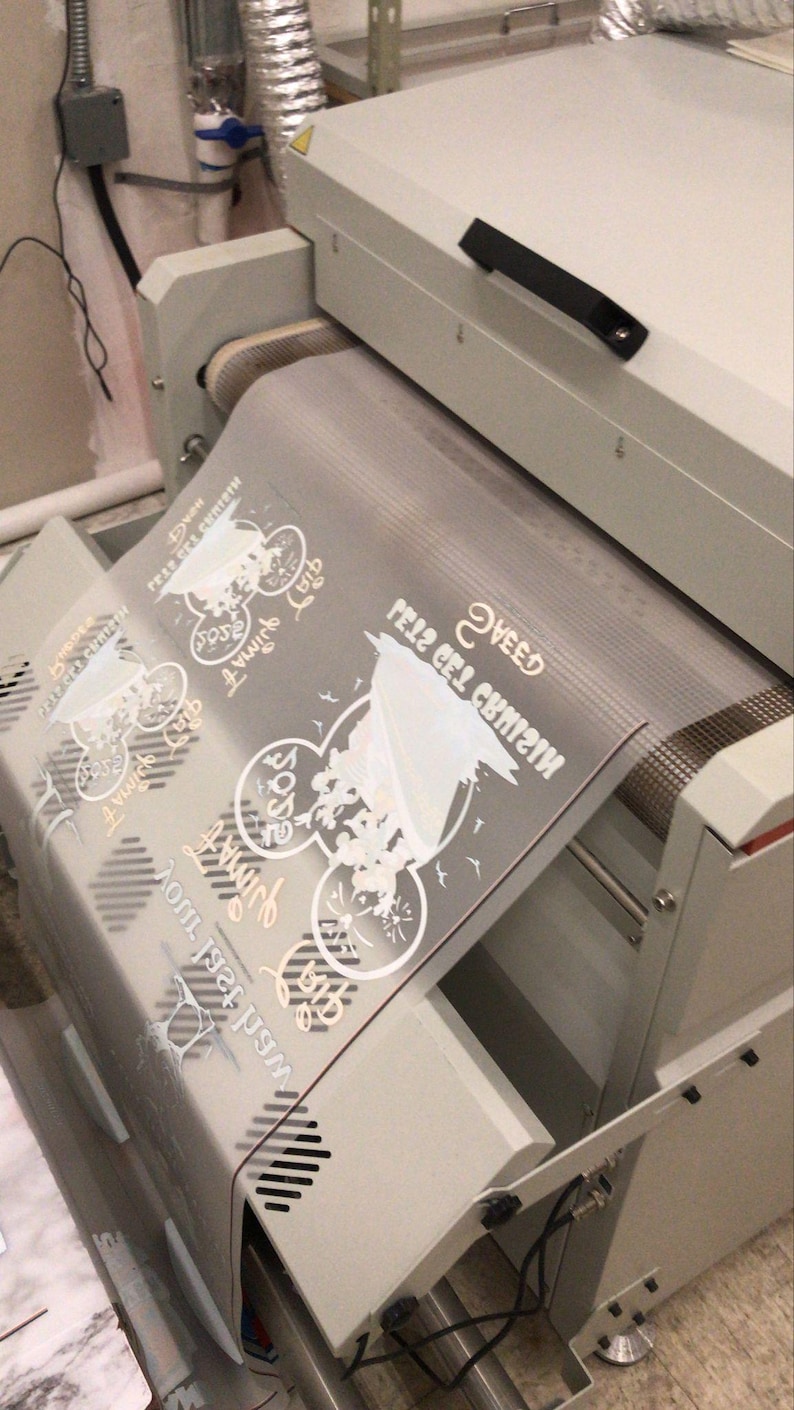 K&ouml;nnte beinhalten: Nahaufnahme einer wei&szlig;en Hei&szlig;presse mit einer durchsichtigen Plastikfolie, auf der ein Design aufgebracht ist. Das Design ist eine wei&szlig;e Silhouette einer Comicfigur mit dem Text "Sweet Feliz del Cumple" auf der Folie.