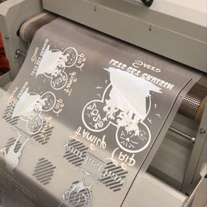 May include: A white heat press machine with a clear plastic sheet on the roller. The sheet has a design printed on it with the text "Getz Get Crazy" and "We're 1st Family".
