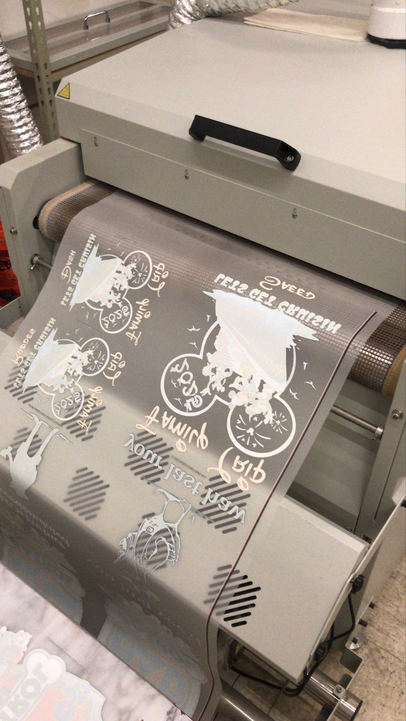 May include: A white heat press machine with a clear plastic sheet on the roller. The sheet has a design printed on it with the text "Getz Get Crazy" and "We're 1st Family".