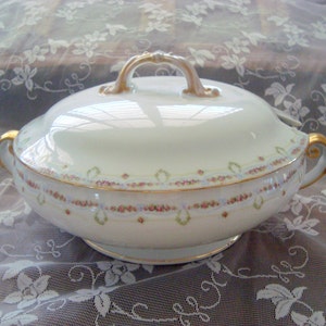 May include: A white porcelain serving dish with a lid, featuring gold accents on the handles and rim. The dish is decorated with a delicate floral pattern in pink, green, and blue, adding a touch of elegance to the design.