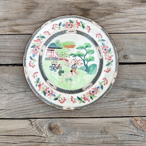 May include: A round, decorative plate with a detailed Asian-inspired design. The plate features a central scene with figures, trees, and a building, surrounded by a floral border in shades of pink, green, and black. The plate is set against a wooden background.