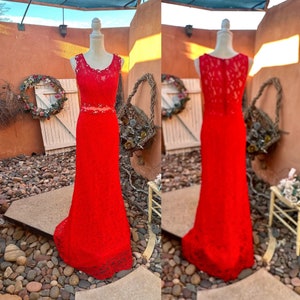 May include: A red lace maxi dress with a fitted bodice and a flowing skirt. The dress has a scoop neckline and a sleeveless design. The dress is displayed on a mannequin.