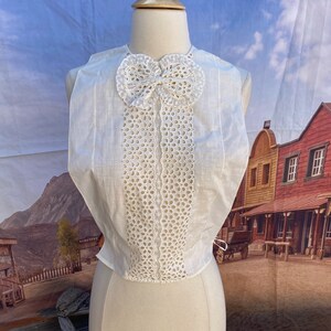 May include: A white sleeveless top with a lace overlay and a large bow at the neckline. The lace is a delicate pattern of circles and the bow is tied in a simple knot.