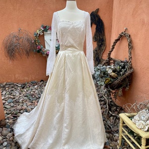 May include: A vintage ivory wedding dress with long lace sleeves and a satin skirt. The dress has a square neckline and is adorned with delicate beading.