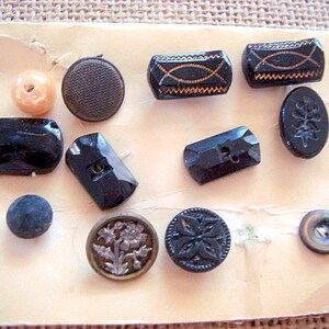 May include: A collection of vintage buttons in various colours and designs, including black, brown, and gold. The buttons are arranged on a white card with a beige background.