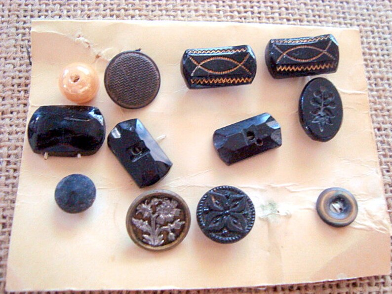 May include: A collection of vintage buttons in various colours and designs, including black, brown, and gold. The buttons are arranged on a white card with a beige background.