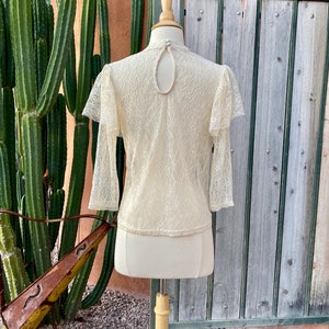 May include: A cream-coloured lace top with long sleeves and a keyhole back. The top has a delicate floral pattern and a ruffled neckline.