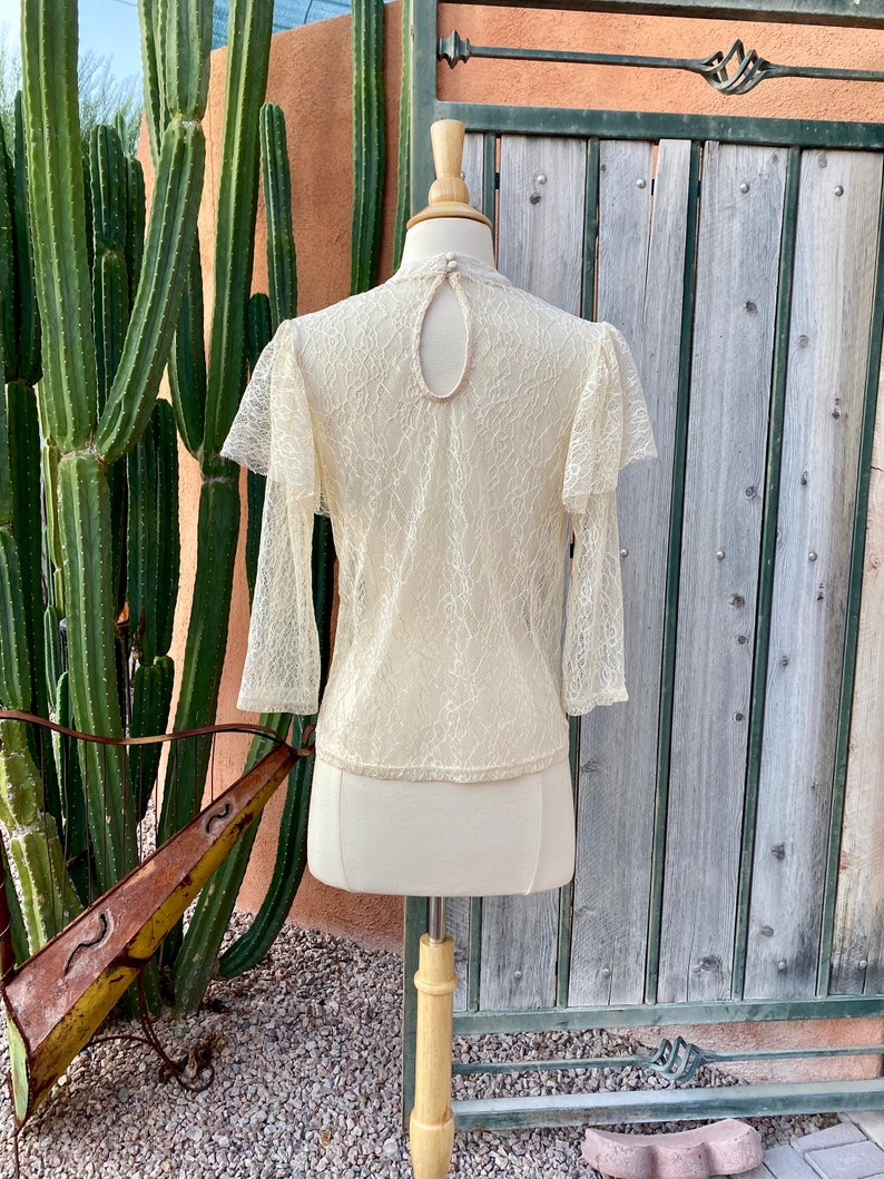 May include: A cream-coloured lace top with long sleeves and a keyhole back. The top has a delicate floral pattern and a ruffled neckline.