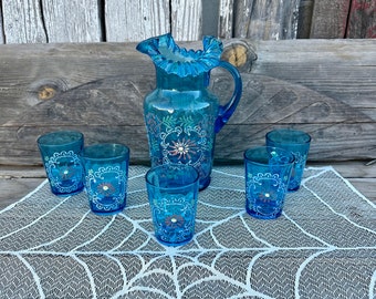 May include: A set of six blue glass cups and a pitcher with a ruffled rim. The glasses and pitcher are decorated with white floral designs. The set is displayed on a white lace tablecloth with a spiderweb pattern.