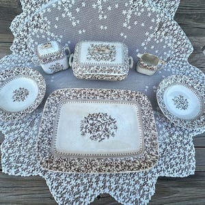 May include: A collection of antique ceramic tableware with a floral pattern in brown and cream. The set includes a rectangular platter, two plates, a covered dish, a creamer, and a sugar bowl, all arranged on a lace tablecloth.