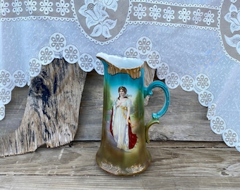 May include: Vintage ceramic jug featuring a hand-painted portrait of a woman in a white gown and red cape. The jug has a teal handle and a gold base. The background depicts a landscape scene.
