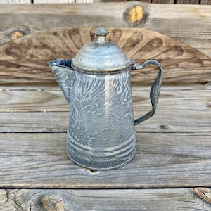 May include: Vintage gray and white speckled enamelware coffee pot with a lid and handle. The pot has a spout for pouring and is sitting on a wooden surface. The coffee pot is a classic kitchenware item.