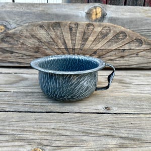 May include: A vintage, blue and white enamelware chamber pot with a handle. The pot has a distinctive marbled pattern and is set against a rustic wooden background. A decorative wooden element is behind the pot.