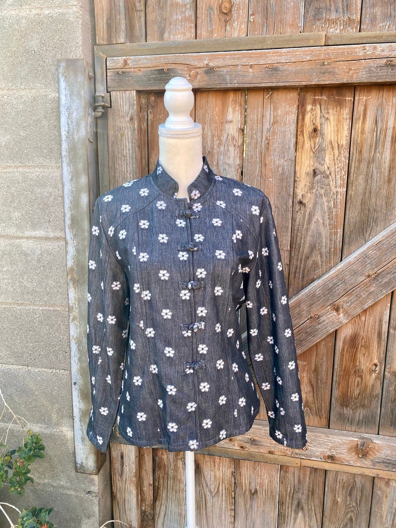 May include: A black denim jacket with white floral embroidery and a mandarin collar. The jacket has five decorative buttons down the front.
