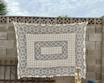 May include: A white crocheted tablecloth with a scalloped edge and a geometric pattern of squares and diamonds. The tablecloth is hanging on a gray brick wall.