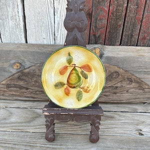 May include: A small, yellow ceramic plate with a painted pear design. The plate is on a wooden easel.