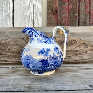 May include: A blue and white porcelain pitcher with a Japanese-inspired design. The pitcher has a rounded body and a curved handle. The design features a landscape with buildings, trees, and water.