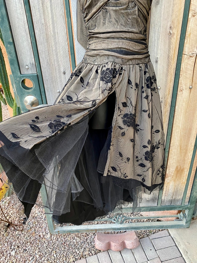 May include: A black and beige tulle dress with a floral pattern. The dress has a fitted bodice and a full skirt. The skirt has a slit in the front and is layered with tulle.