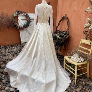 May include: A vintage ivory satin wedding dress with long sleeves and a lace bodice. The dress has a full skirt and a train. The dress is on a mannequin in a garden setting.