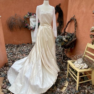 May include: A vintage ivory wedding dress with long sleeves and a lace bodice. The dress has a flowing skirt and is displayed on a mannequin in a rustic setting.