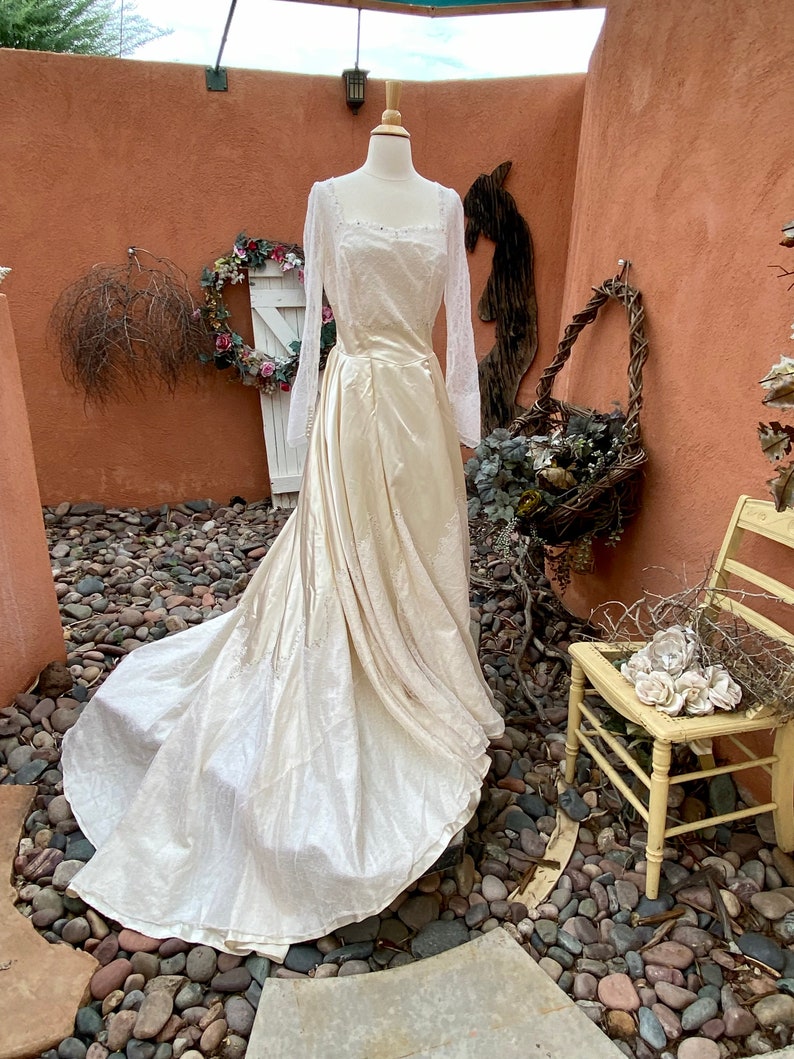 May include: A vintage ivory wedding dress with long sleeves and a lace bodice. The dress has a flowing skirt and is displayed on a mannequin in a rustic setting.