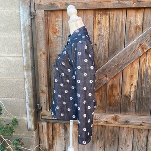 May include: A black denim shirt with a white floral pattern. The shirt has a mandarin collar and long sleeves. It is displayed on a white mannequin against a wooden background.