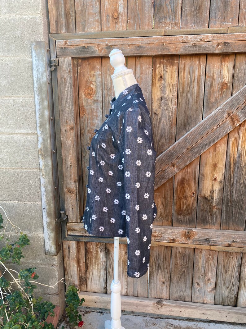 May include: A black denim shirt with a white floral pattern. The shirt has a mandarin collar and long sleeves. It is displayed on a white mannequin against a wooden background.